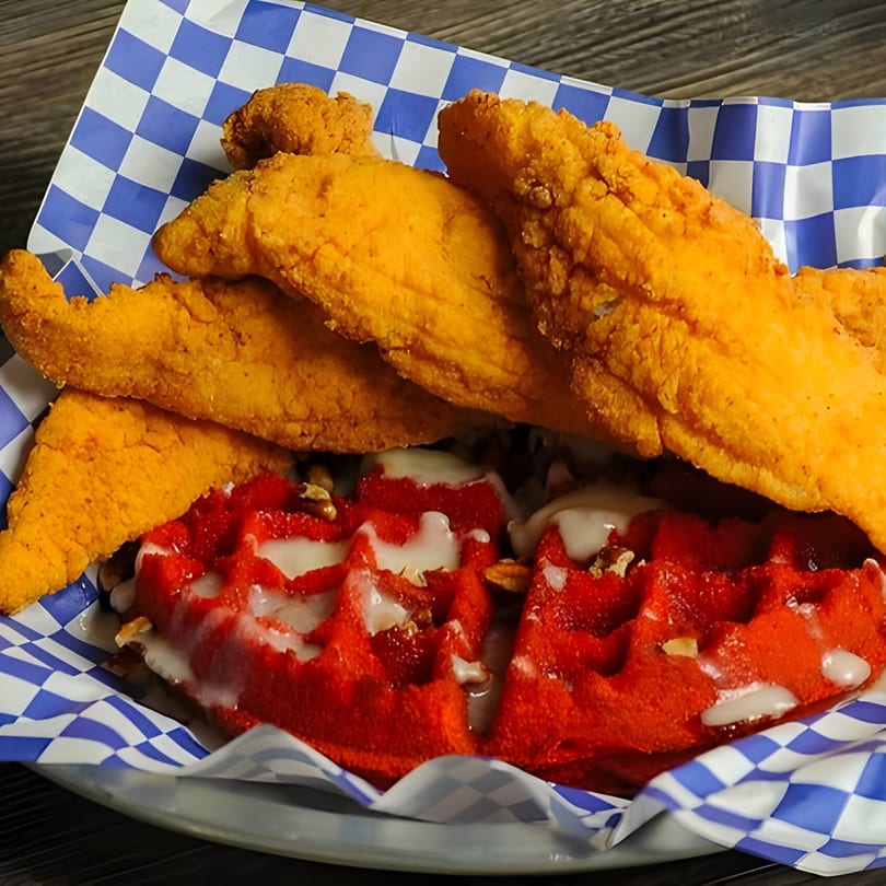 Crispy fried fish fillets and red velvet waffles with icing and nuts in a blue checkered basket.