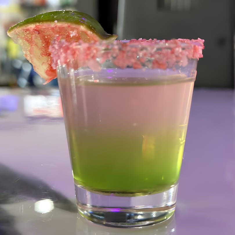 A vibrant pink and green layered cocktail, garnished with a watermelon slice and pink sugar rim.